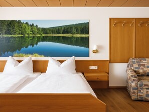 A bedroom with a large bed, a nightstand, a wooden wardrobe, a patterned armchair, and a wall-mounted painting of a lake. - Hotel zur Linde (Lohberg)