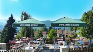 Outdoor seating with tables and chairs, two green umbrellas with text, and a mountainous backdrop. - Hotel zur Linde (Lohberg)