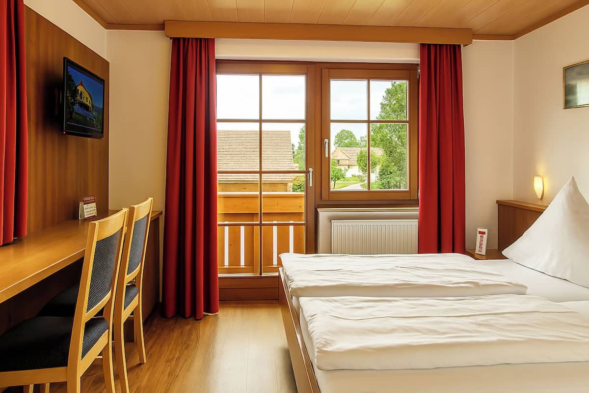 Une chambre d’hôtel avec un bureau en bois, deux chaises, un lit, une fenêtre avec des rideaux et une vue sur l’extérieur.