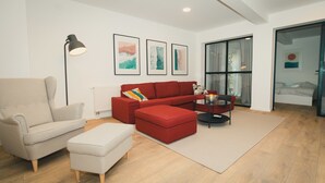 A modern living room with a beige armchair, a red sectional sofa, a small ottoman, a glass coffee table, and a floor lamp. There are three framed artworks on the wall, a large window with sheer curtains, and a bed in the background. - Apartments Franky6 (Zagreb)