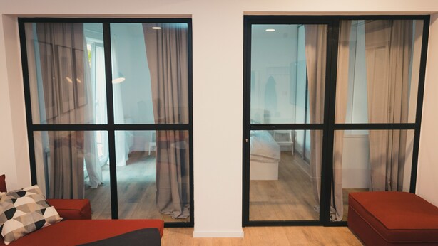 A modern room with a large sliding glass door leading to a hallway. There is a red sofa and a small red ottoman in the room. - Apartments Franky6 (Zagreb)
