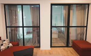 A modern room with a large sliding glass door leading to a hallway. There is a red sofa and a small red ottoman in the room. - Apartments Franky6 (Zagreb)