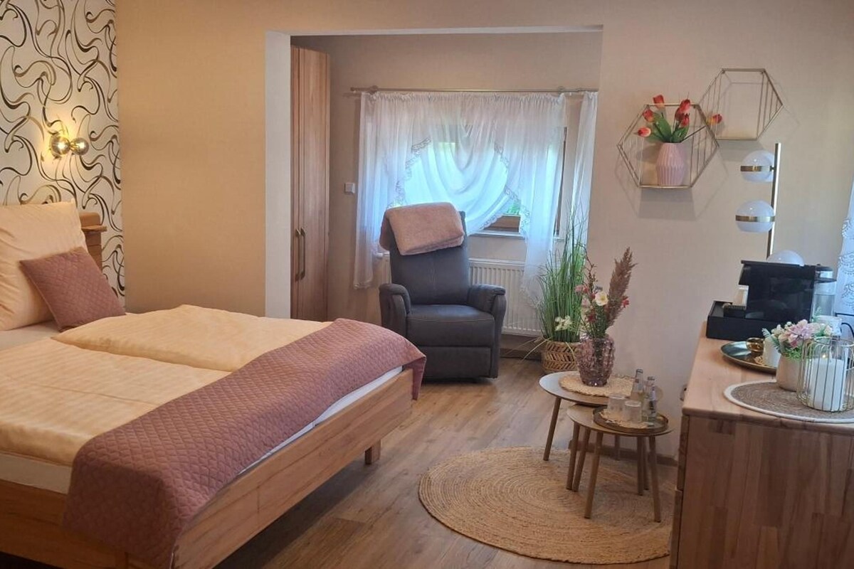 A hotel room with a bed, a chair, a small table, and a desk with a lamp.