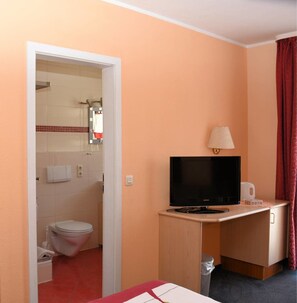Habitación de hotel compacta con baño visible a través de una puerta abierta, un escritorio con televisor, lámpara y un pequeño cubo de basura.