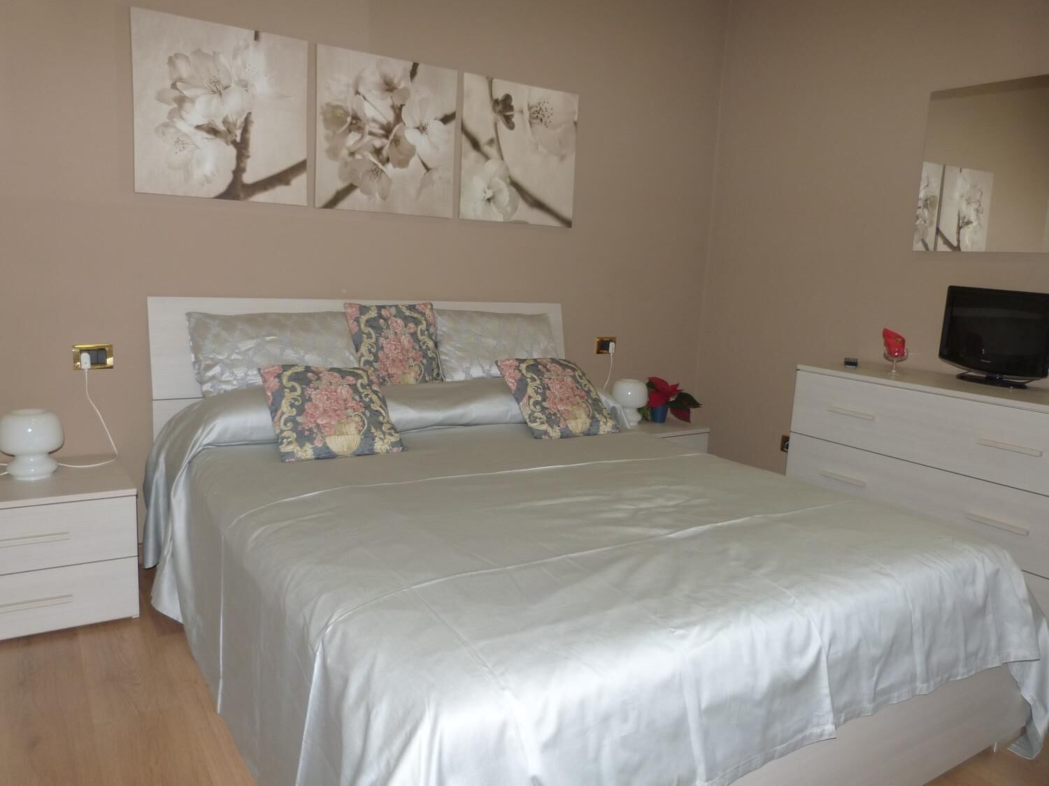 Un dormitorio con cama, mesitas de noche, cómoda, televisor y decoración en la pared.