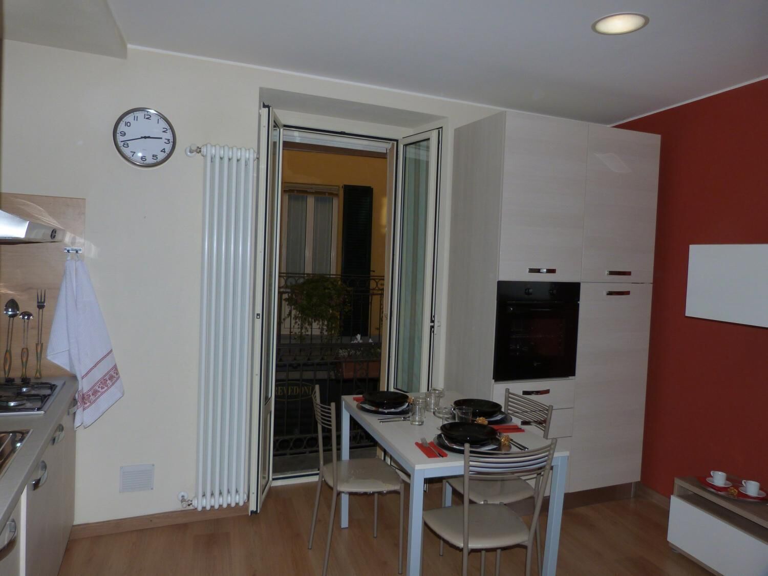 A compact kitchen with a dining table set for two, a clock on the wall, and a TV mounted above the cabinets.