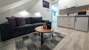 A modern living room with a dark grey sofa, a round wooden coffee table, and a patterned rug. A small table lamp with a geometric design is on the coffee table. The room has a sloped ceiling and a kitchen area with white cabinets and a microwave. - Apartments Beautiful You Are 2 (Zagreb)