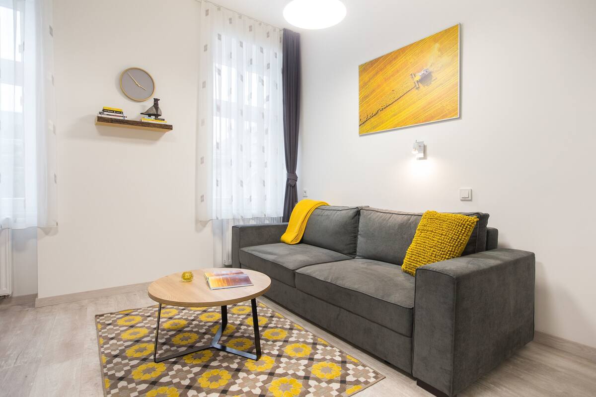 A living room with a grey sofa, a yellow throw pillow, a round wooden table, a patterned rug, a wall clock, a shelf with books, and a painting.