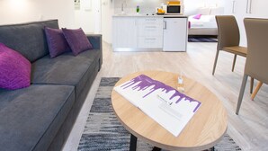 A modern living room with a grey sofa, a wooden coffee table with a document, a kitchen area with a microwave, and a bed with a white headboard. - Apartments Beautiful You Are 2 (Zagreb)