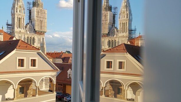 A view through a window of a building with a large, ornate church with twin spires and scaffolding, and adjacent residential buildings with red-tiled roofs and arched windows. - Apartments Beautiful You Are 2 (Zagreb)
