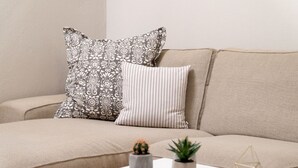 A beige sofa with patterned pillows, a white side table with two potted plants and a geometric decoration. - House Loft Foursquare (Thessaloniki)
