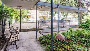 Un área de estar al aire libre con techo, bancos, barandilla metálica y vista a un estacionamiento y edificios al fondo.