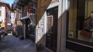 A narrow cobblestone street with traditional European buildings, a shop with a sign, and a large window displaying a mannequin. - Appartamento Il Borgo (Bellagio)