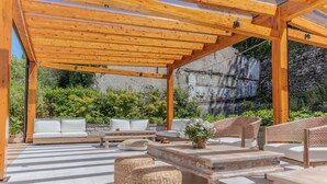 An outdoor patio with wooden pergola, white sofas, wicker chairs, and a stone table. - Appartamento Il Borgo (Bellagio)