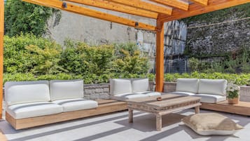 An outdoor patio with a wooden pergola, white cushioned seating, and a stone wall backdrop.