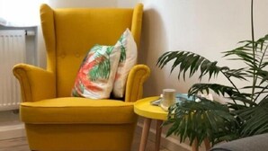 Een gele fauteuil met een enkel decoratief kussen, een klein rond tafeltje met een kopje, en een grote groene plant.