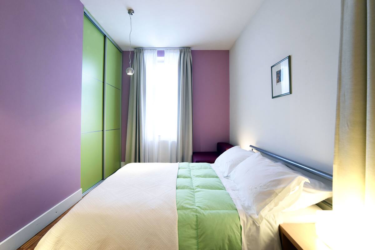 A bedroom with a bed, a window with curtains, a bedside table, and a wall-mounted picture.