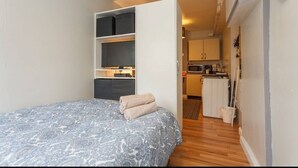 Een compacte slaapkamer met een bed, een inbouwkast en een kitchenette op de achtergrond.