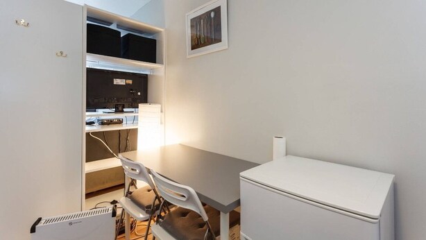 Een compacte kamer met een bureau, stoel en een witte koelkast. Er is een plank met diverse spullen en een lijst aan de muur.