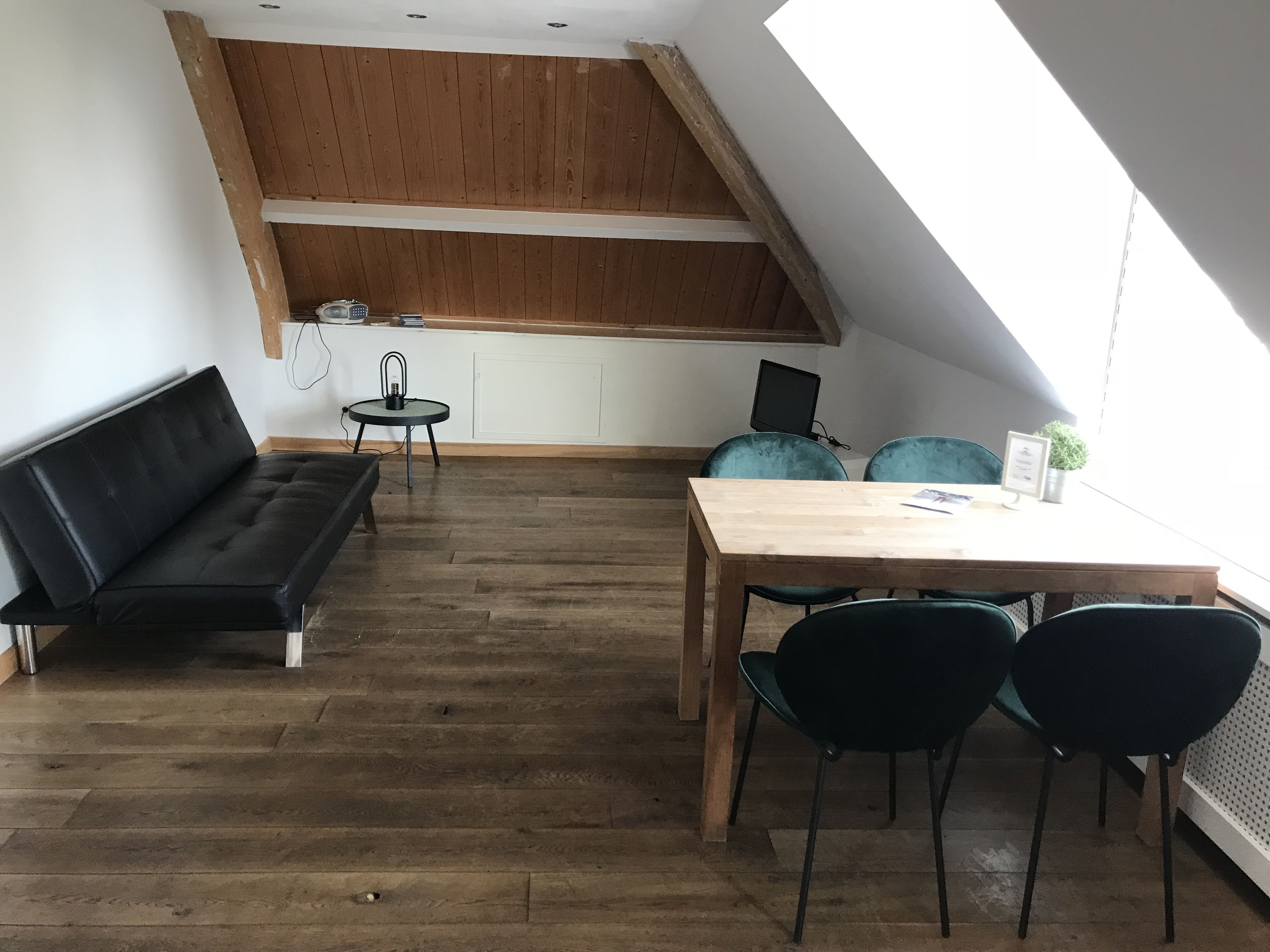 A loft-style room with a wooden floor, a black sofa, a wooden dining table with chairs, a small round table with a lamp, and a laptop on a white desk.