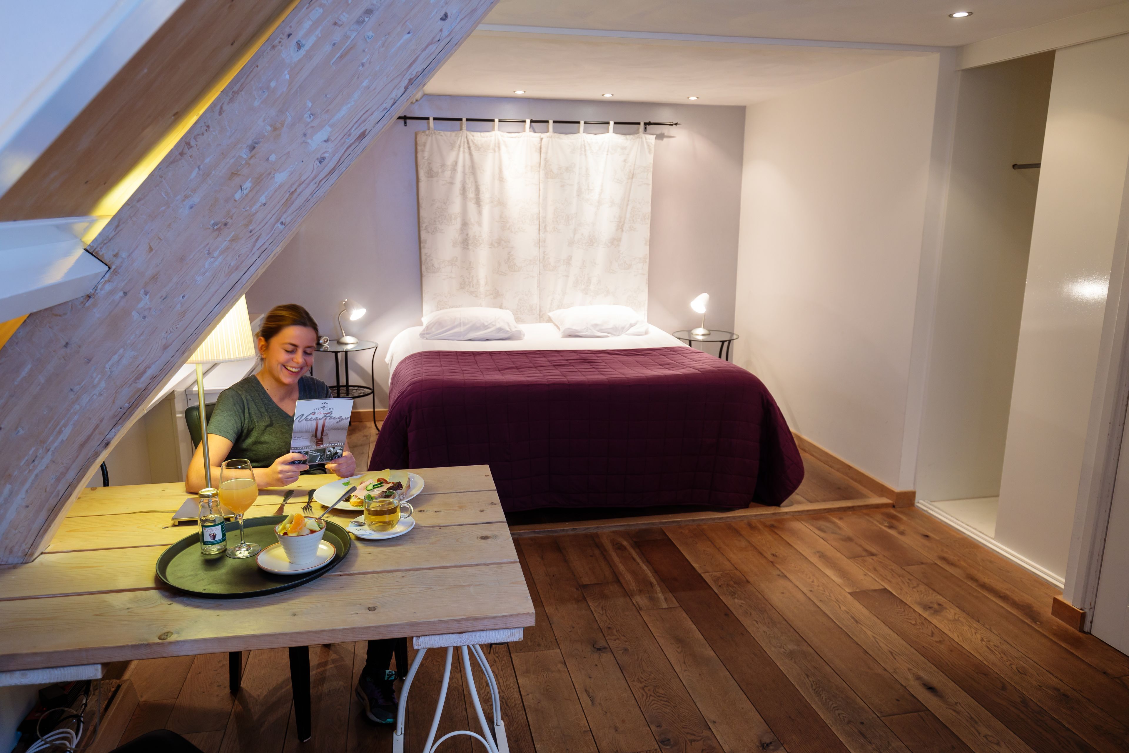 Una habitación pequeña y acogedora con una mesa de madera, una cama con una manta roja y una mesita de noche con una lámpara.