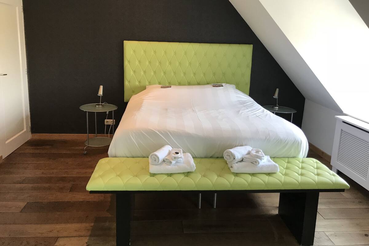 A neatly made bed with a green headboard and footboard, a small bedside table, and a bench with towels.