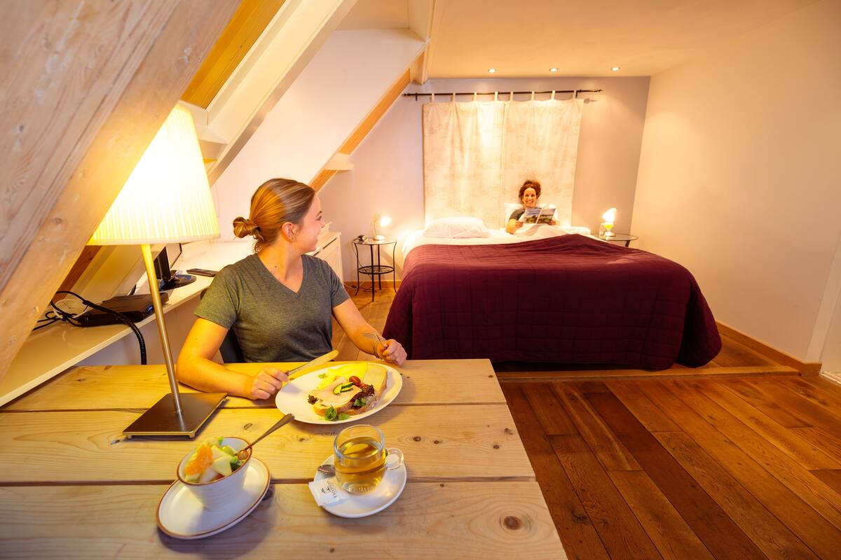 A cozy room with a wooden table, a bed with a red bedspread, and a person reading a book.