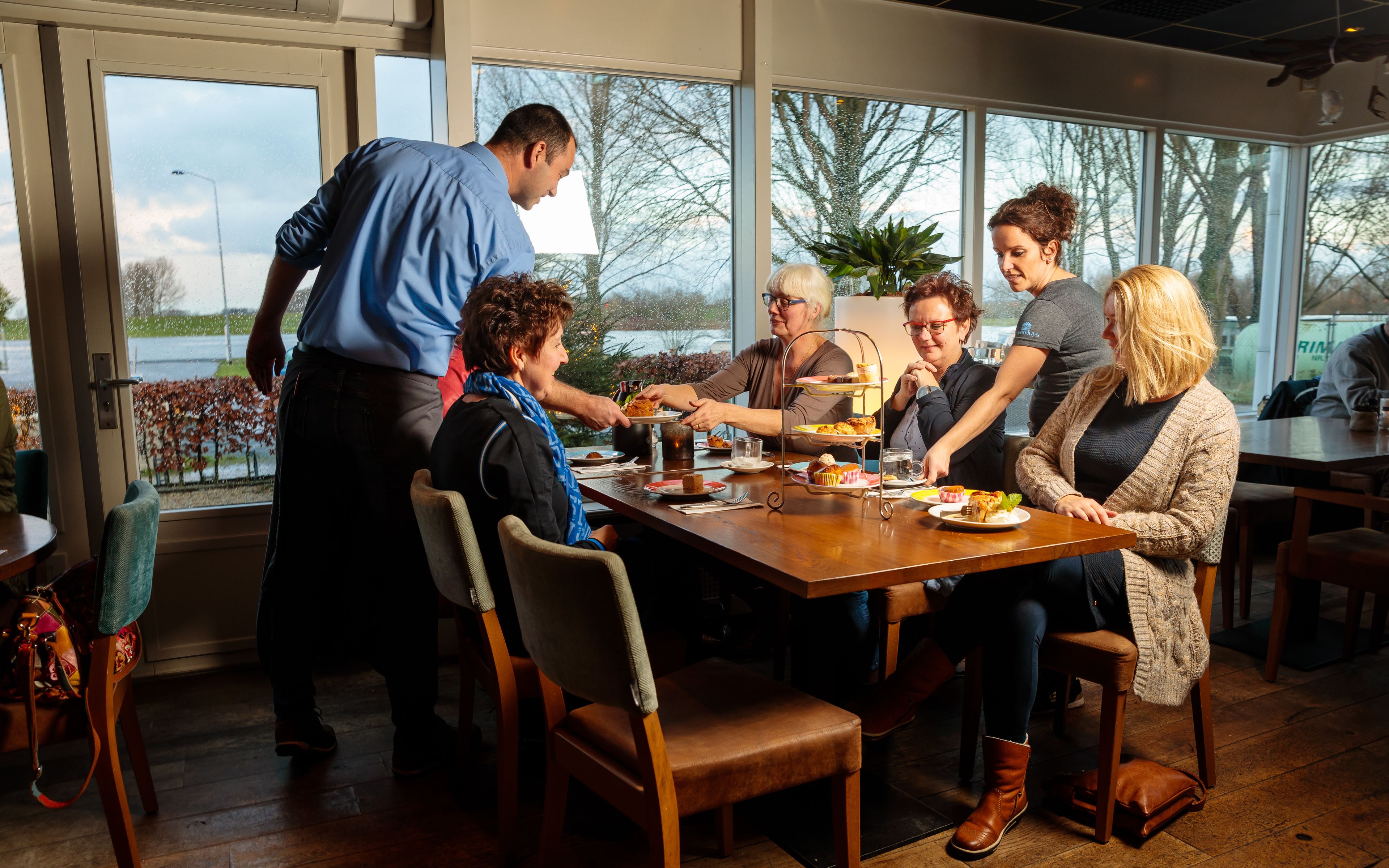 Un grupo de personas sentadas alrededor de una mesa de madera en una habitación con ventanales amplios, disfrutando de una comida.