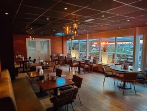 Un restaurante espacioso con suelo de madera, varias mesas y sillas, y ventanas amplias que ofrecen vistas al exterior.