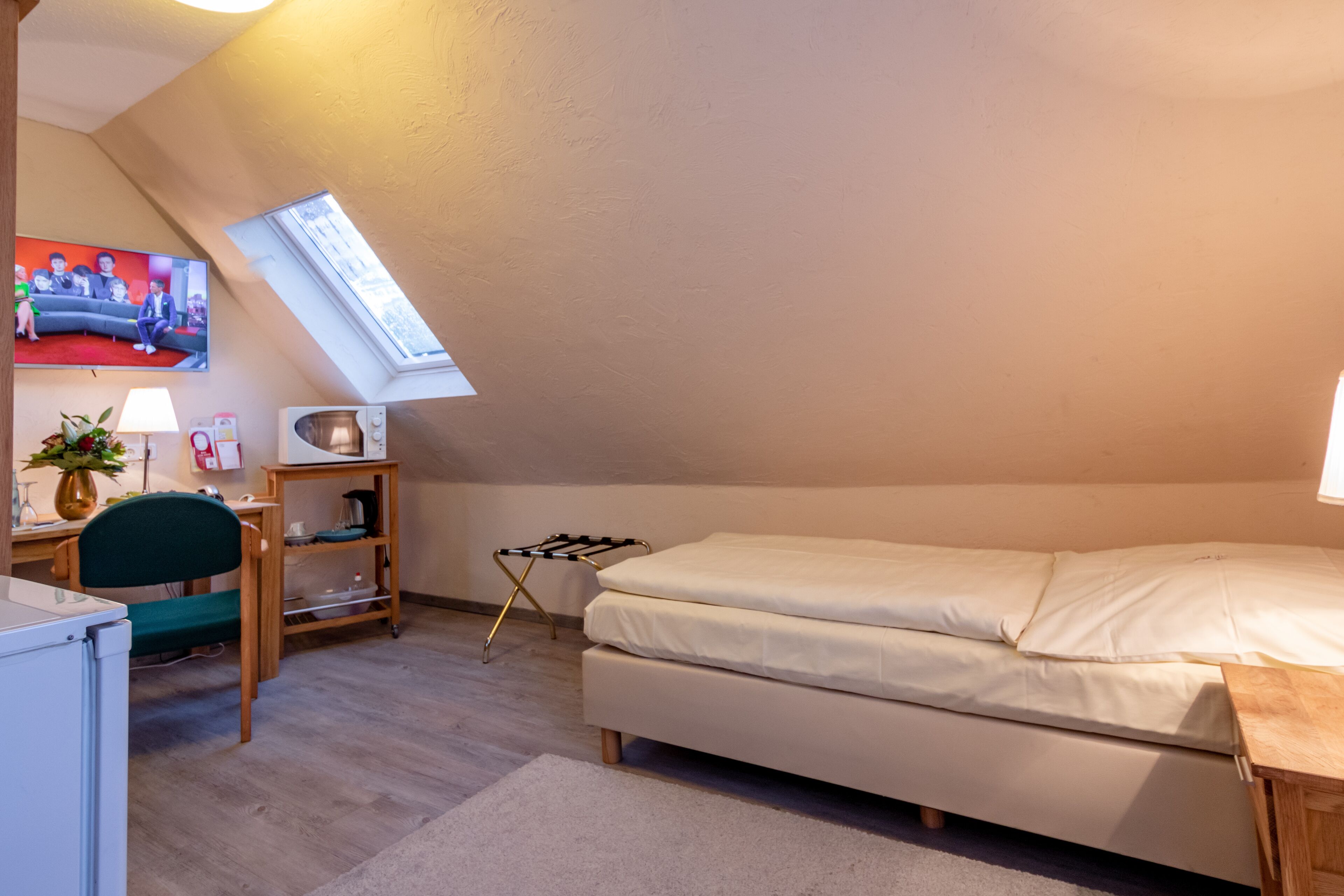 A small, cozy room with a loft bed, a desk with a chair, a TV mounted on the wall, and a skylight.