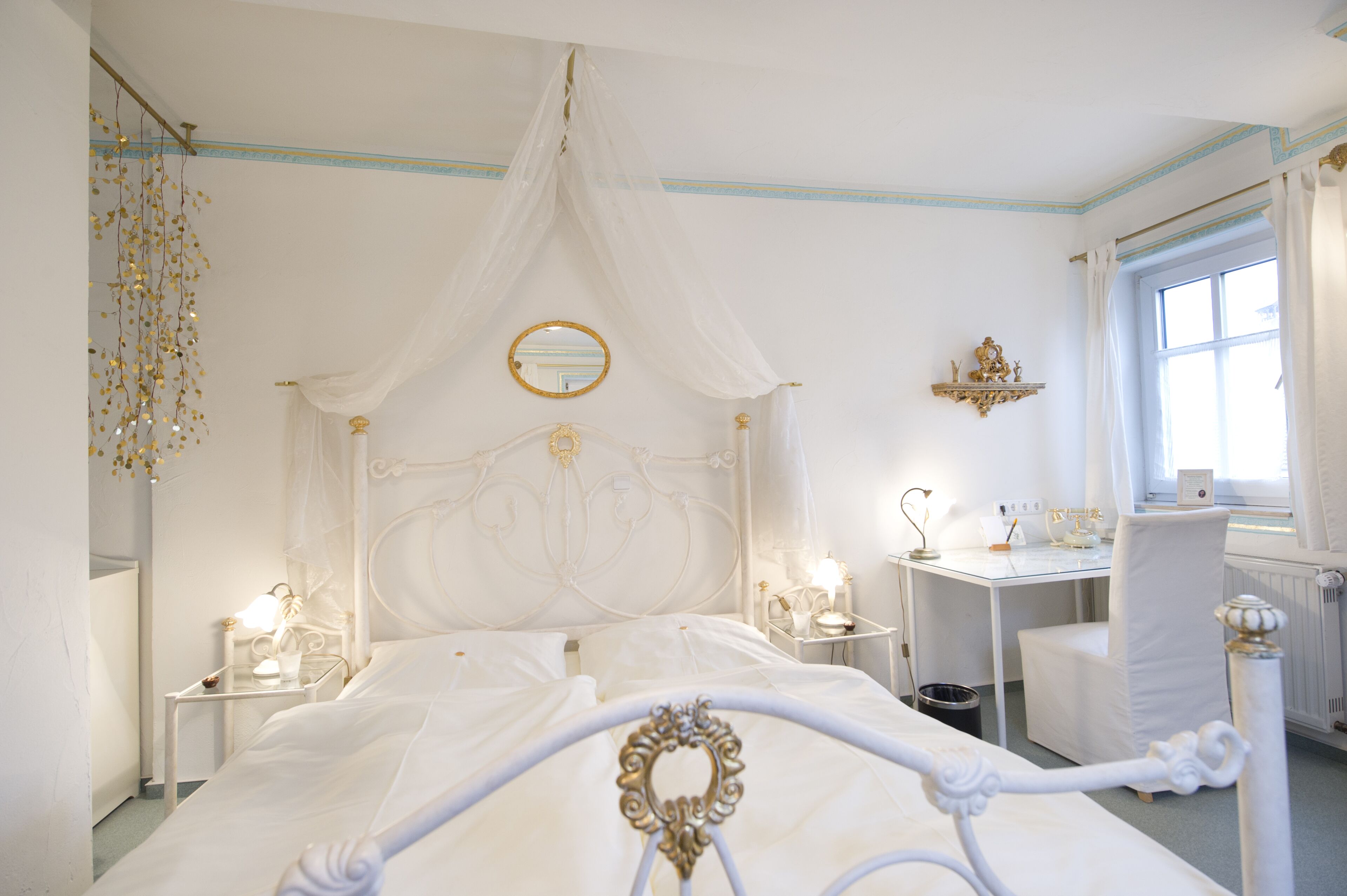 A white four-poster bed with a canopy, bedside tables with lamps, a small desk, and a chair.