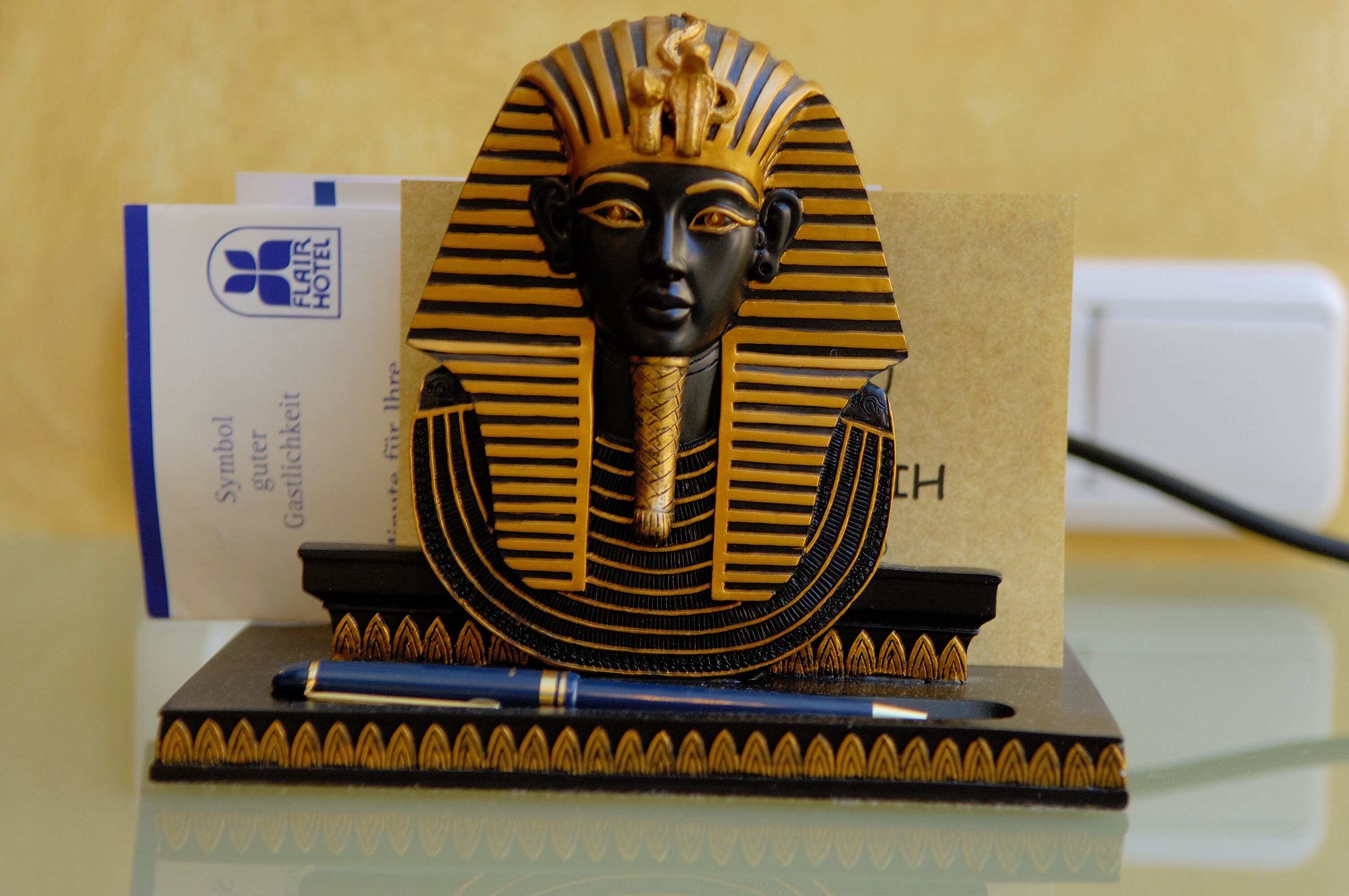 Ein dekoratives Objekt, das an einen antiken ägyptischen Artefakt erinnert, möglicherweise ein Büstenbild, mit goldenem Kopfschmuck und einer Halskette, platziert auf einer spiegelnden Oberfläche.