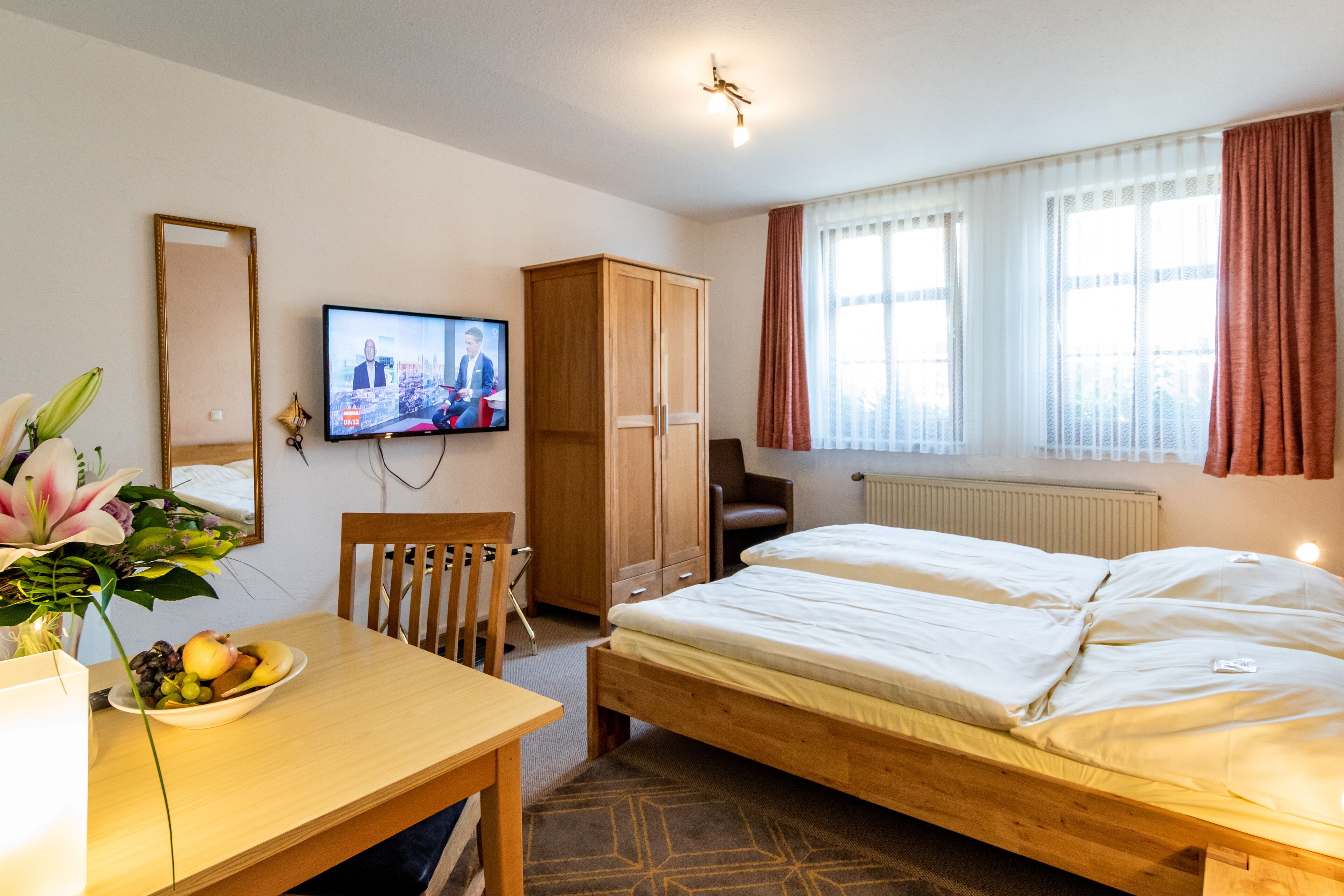 Habitación de hotel con una cama de madera, un escritorio con frutero, televisor, armario y ventana con cortinas.