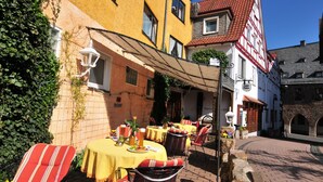 An outdoor café with tables and chairs, a yellow awning, and a cobblestone street. - Hotel Goldflair am Rathaus (Korbach)