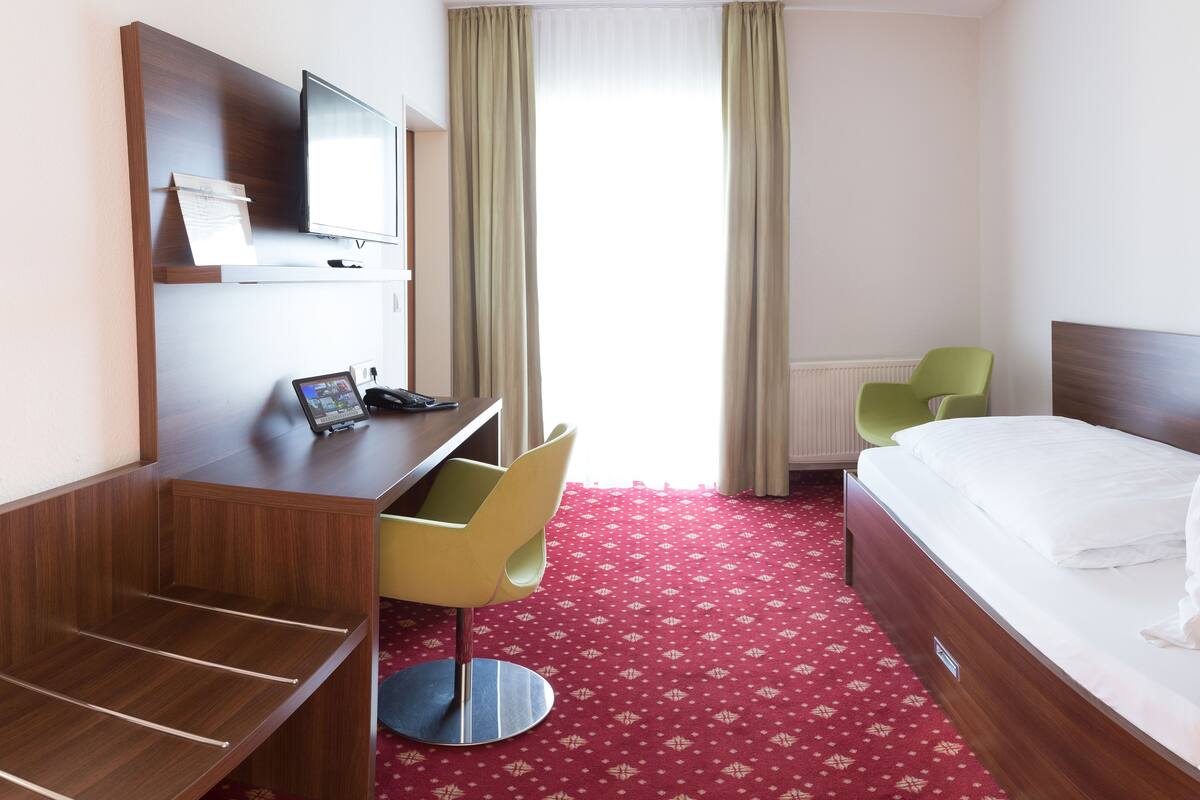 A hotel room with a desk, chair, bed, and a view of the outdoors through a window with curtains.