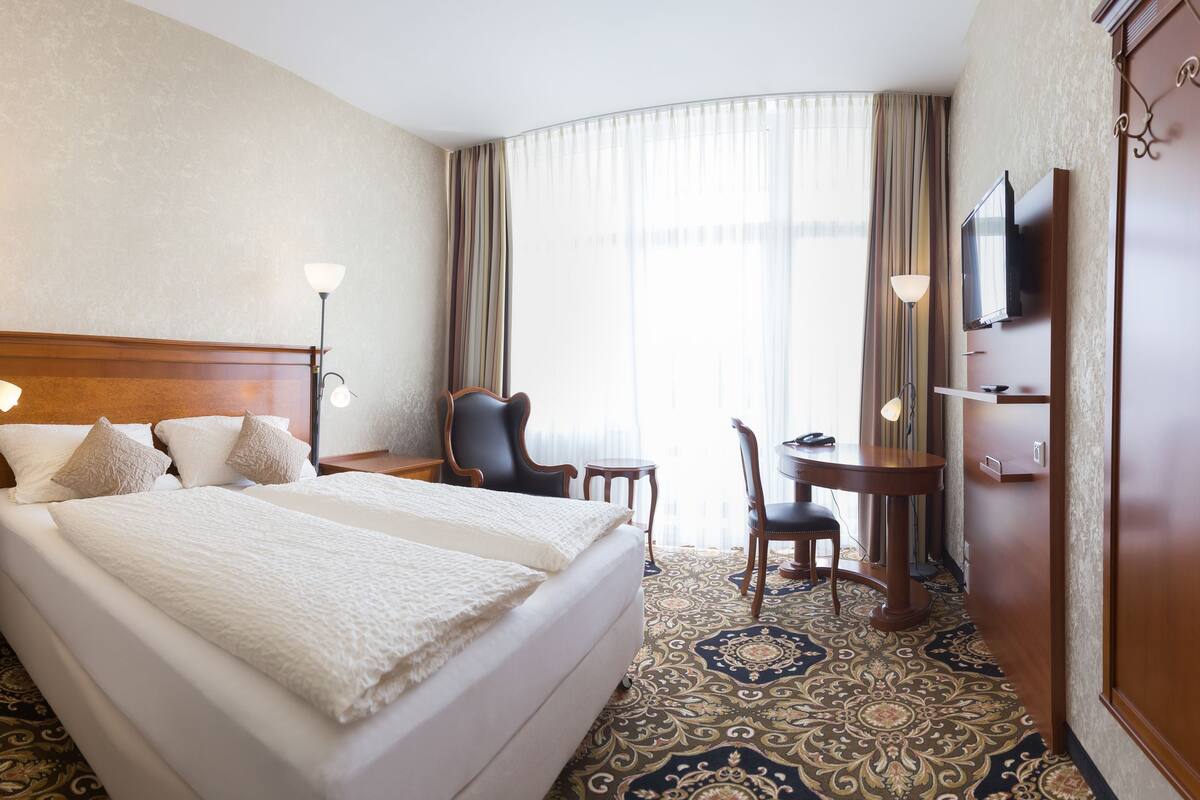 A hotel room with a large bed, bedside tables, a desk with a chair, a television, and a large window with curtains.