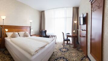 A hotel room with a large bed, bedside tables, a desk with a chair, a television, and a large window with curtains.
