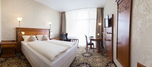 A hotel room with a large bed, bedside tables, a desk with a chair, a television, and a large window with curtains. - Vitalhotel König am Park (Bad Mergentheim)
