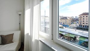 Une chambre avec un lit, une fenêtre donnant sur des immeubles et une tour, et une lampe.