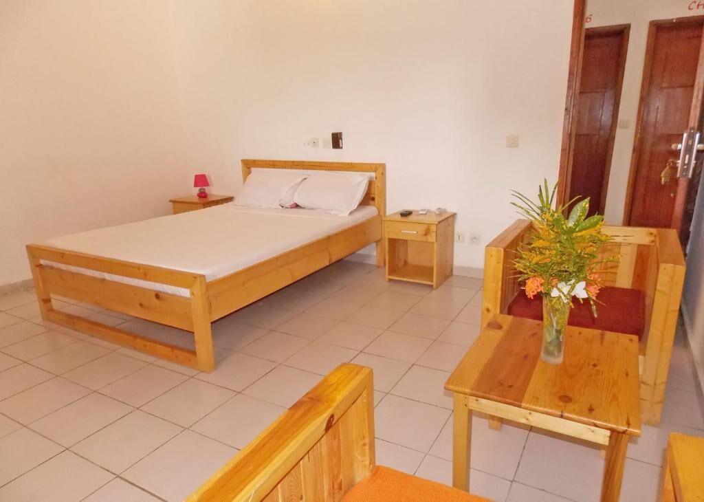 Una habitación con una cama de madera, una mesita de noche pequeña con una lámpara, una mesa de madera con un jarrón de flores y una silla de madera.