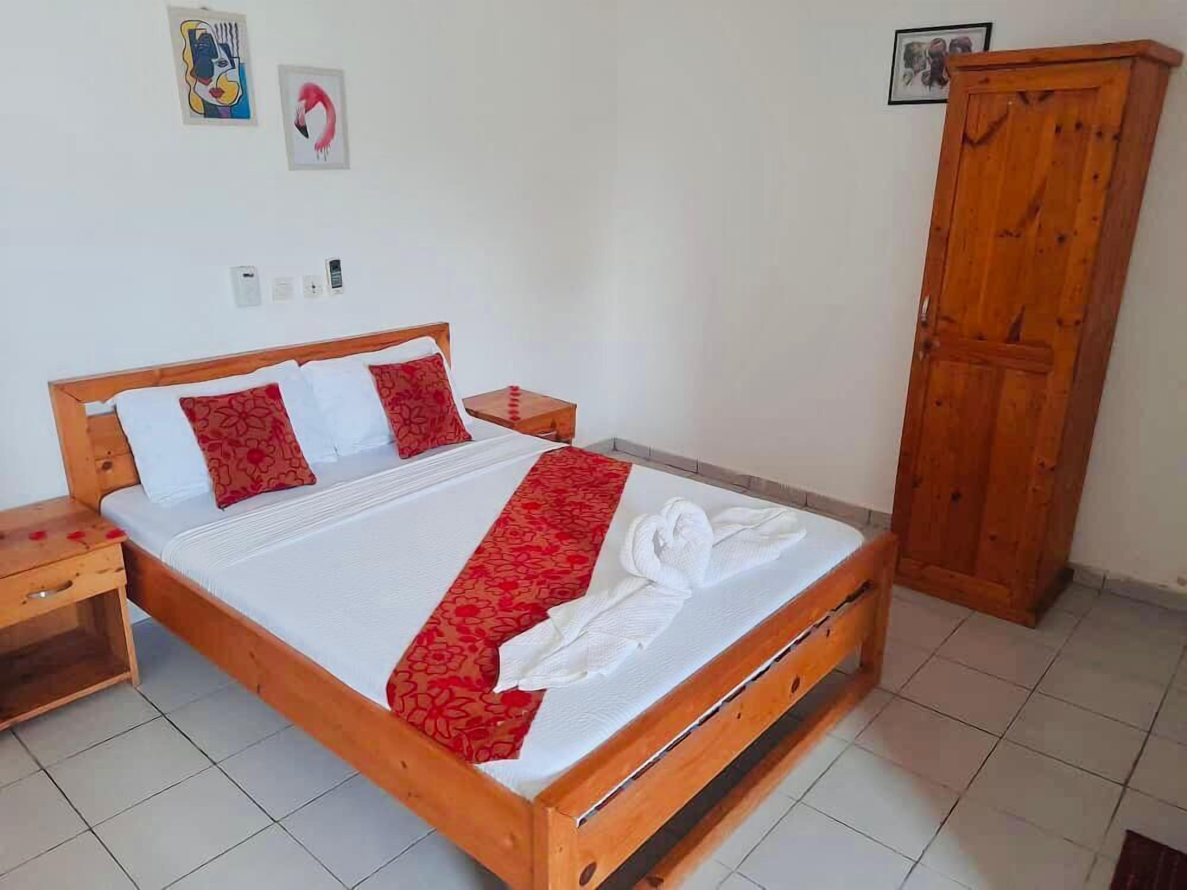 Una cama de madera con ropa de cama blanca y roja, una mesita de noche de madera, un armario de madera y una pared con dos cuadros enmarcados.