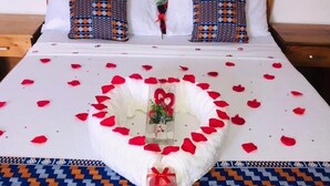 A neatly made bed with a heart-shaped arrangement of rose petals and a small gift box, surrounded by patterned bed runners. - Imans Hotel Dabou (Dabou)