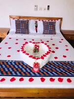 Una cama bien hecha con un arreglo de pétalos de rosa en forma de corazón y una pequeña caja de regalo, rodeada de corredores de cama estampados.