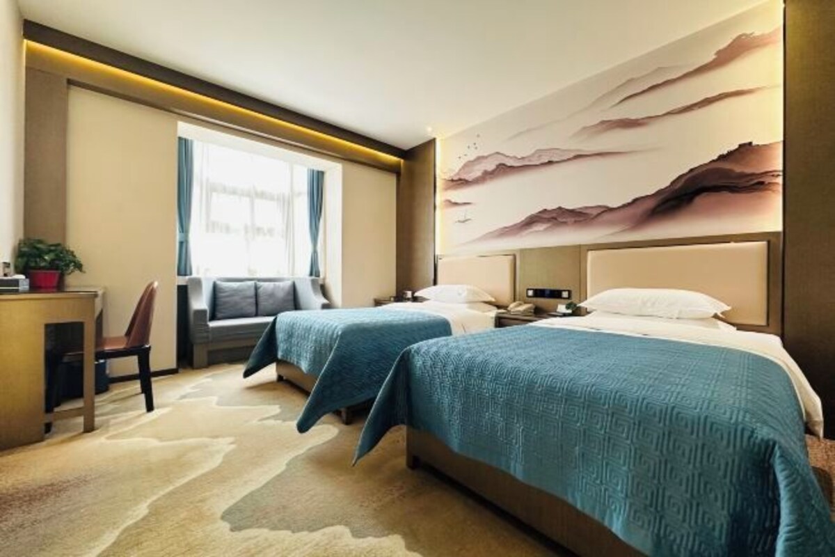 A hotel room with two beds, a desk, a chair, a window with curtains, and a wall with a textured mural.