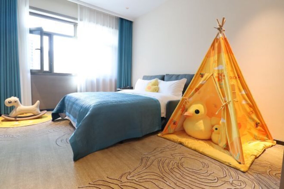 A hotel room with two beds, a window with curtains, a rocking horse, and a teepee with stuffed animals.