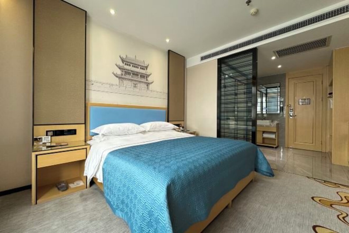 A hotel room with a bed, a nightstand, a desk, a wardrobe, and a wall with a drawing.