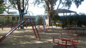 Ein Spielplatz mit verschiedenen Spielgeräten, darunter eine Rutsche und Schaukeln, umgeben von Bäumen und einem überdachten Sitzbereich.