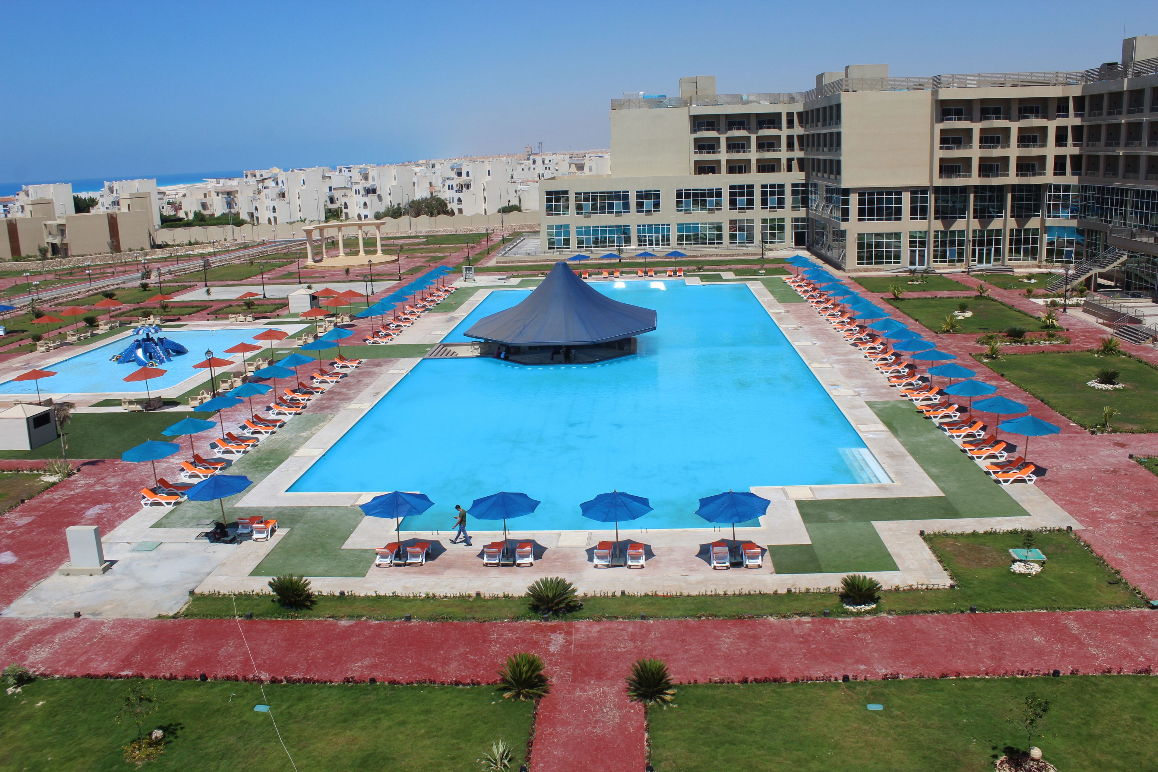 A large swimming pool with a central water slide, surrounded by sun loungers and umbrellas, with a view of residential buildings in the background.
