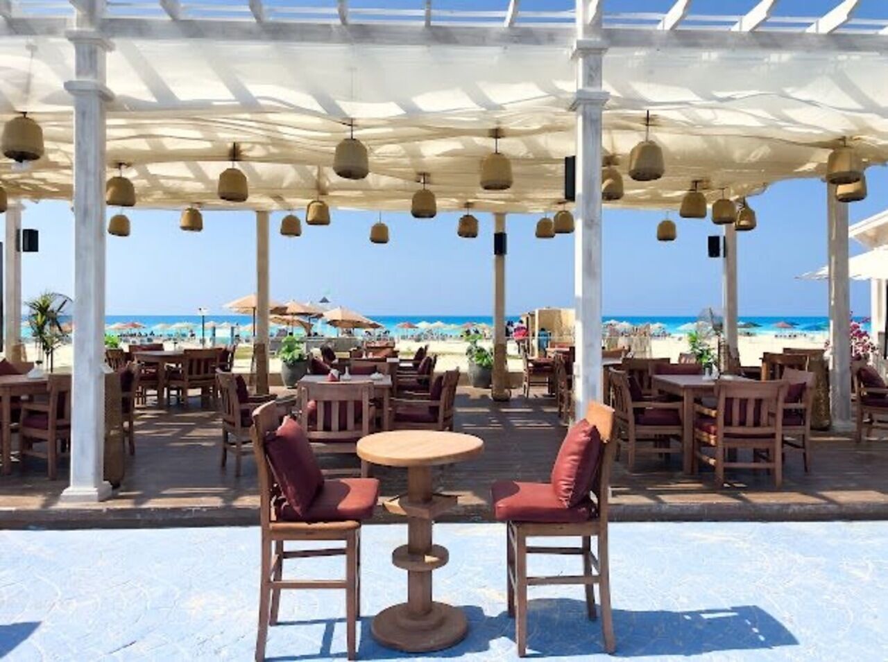 Ein Sitzbereich im Freien mit Tischen und Stühlen, eine Pergola mit hängenden Lichtern und Blick auf den Strand.