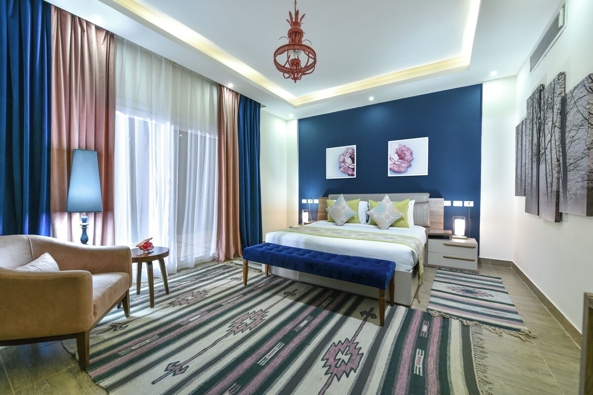 Una camera d'albergo con un letto, una poltrona, un comodino, una lampada, un lampadario e opere d'arte alle pareti.